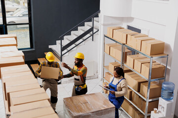 Package handlers team lifting heavy parcel to prepare for transportation. African american warehouse loader standing on ladder and taking cardboard box from shelf top view