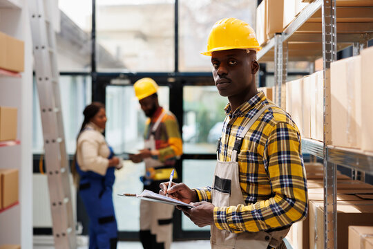 African American Man Writing On Clipboard In Industrial Warehouse Portrait. Post Office Storehouse Package Handler Wearing Helmet Using Orders Checklist And Looking At Camera