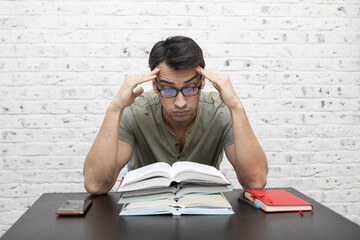 Upset young male student covers his face with hand and sits at the table with a lot of books....
