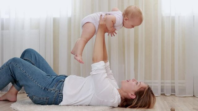 Happy loving millennial caucasian mom lie on back on floor in light cozy bedroom lift up toss in air small baby daughter or son.Caring mum laugh cuddle kiss cute infant babe have fun enjoy motherhood.