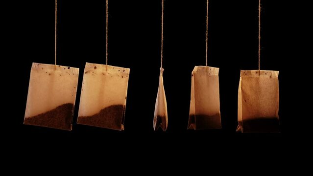 Collection of used brown paper tea bags with tasty herbal brew hang swaying on thin threads on black background close view