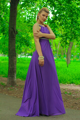 blonde beautiful natural girl with blonde hair in a lilac dress in nature, in the park, in the garden by a tree, with makeup and hairstyle without a bra