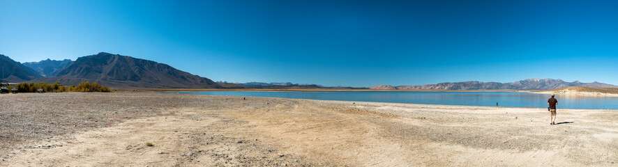 Obraz premium Panoramic view of Crowley Lake and the surrounding mountains, during a sunny day. A person walks near the edge of the lake.
