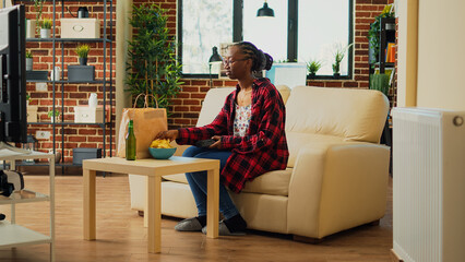 African american girl receiving takeaway meal at home, preparing to watch movie on television and eat fast food in paperbag. Young cheerful woman enjoying film on tv, drinking beer.