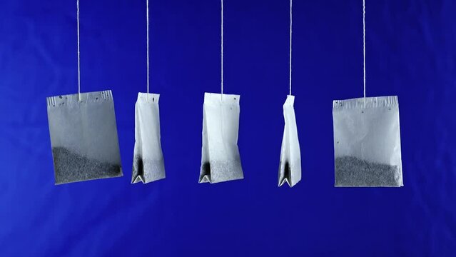 Line of new white paper tea bags with tasty natural brew hang turning on thin threads on deep blue background close view