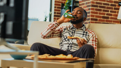 African american guy eating pizza at television and laughing in living room Happy man enjoying delivery food and watching favorite action movie on tv, having fun at home. Handheld shot.