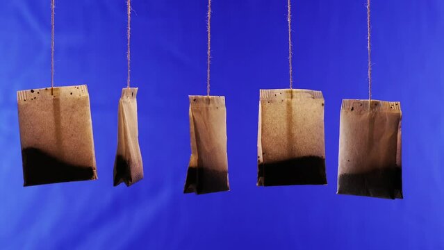 Group of used brown paper tea bags with tasty herbal brew hang turning on thin threads on deep blue background close view
