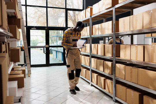 Warehouse Supervisor Doing Freight Revision, Taking Notes And Listening To Music In Headphones. African American Young Shipment Department Operator Supervising Storage Merchandise