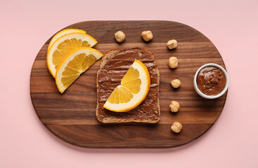 Wooden board of tasty toast with hazelnut butter and orange slices on pink background