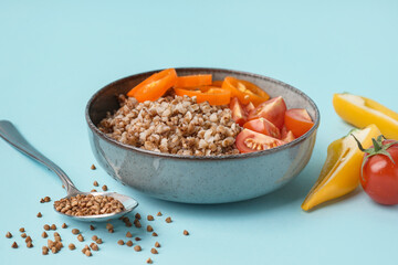 Bowl of tasty buckwheat porridge with fresh vegetables on blue background