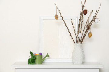 Vase with willow branches, Easter eggs, rabbit and painting on shelf near light wall