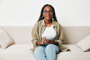 African American woman business freelancer working sitting on the couch at home in the phone, business calls and messages happiness smile, home clothes and eyeglasses, light interior background.