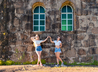 Obraz na płótnie Canvas Female traveler friends posing for a photo and having fun in front of an old stone wall with two green windows