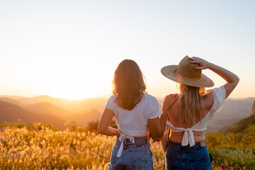 Two female friends standing next to each other watching the sunset in the background. Young woman...