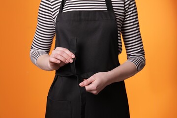 Woman putting on black apron against orange background, closeup - Powered by Adobe
