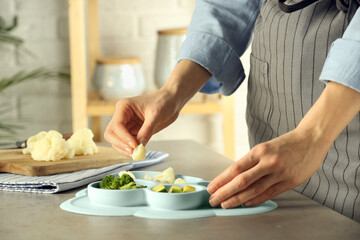 Baby food. Woman putting piece of cauliflower into section plate with different vegetables at grey textured table in kitchen, closeup