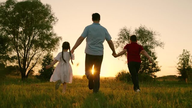 Father, son, daughter play, run on green grass, hold hands in park. In summer, family walks outdoors holding hands. Family Teamwork. Weekend with family. Silhouette, happy running kids, dad, nature.