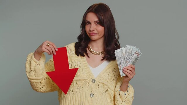 Upset pretty woman showing red arrow pointing down, concept of downgrade, unsuccessful business, fall of stock market money dollar exchange rate bankruptcy fail. Young adult girl on gray background