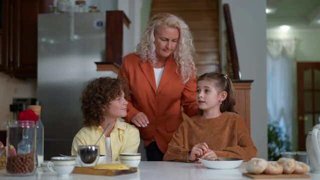 Portrait of happy loving mother talking with children sitting at table with chocolate cereal. Smiling Caucasian woman enjoying morning with son and daughter at home indoors