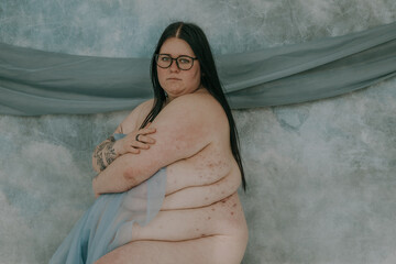 fat woman with glasses and black hair sitting hugging her chest looking at camera