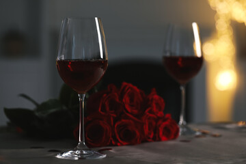 Glasses of red wine and rose flowers on grey table against blurred lights, space for text. Romantic atmosphere