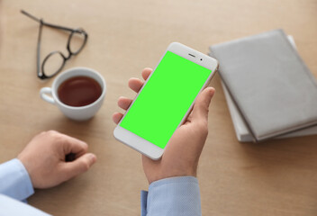 Man holding smartphone with green screen at wooden table indoors, closeup. Gadget display with chroma key. Mockup for design