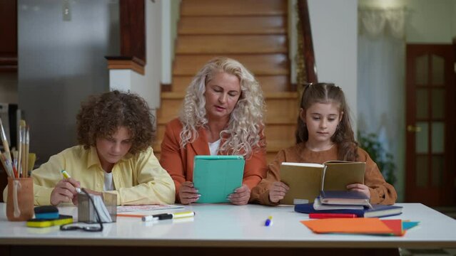 Concentrated positive mother surfing Internet on digital tablet watching son and daughter doing homework. Portrait of intelligent busy Caucasian woman helping children with studies at home