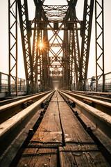 Train tracks lead downwards over a long metal bridge. There is a warm evening atmosphere.