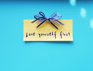 Yellow note with ribbon on blue wall with handwritten text  LOVE YOURSELF FIRST, concept of self talk affirmation, put yourself as first priority with self love and self respect