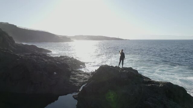 aerial shot of a young woman in orbit that is on a rock near the sea and waves. In the city of Galdar, Gran Canaria.