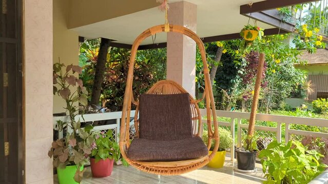 a hanging chair swing made of wood swinging in the wind in the veranda surrounded by plants and the green garden