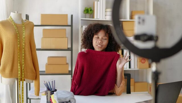 Female African designer and online seller is live streaming broadcast for selling new clothes and receiving the order from customer  and packaging product in workspace.