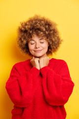 Portrait of kind lovely curly-haired European woman holding hands in prayer with closed eyes in a wish indoor studio shot isolated over yellow background