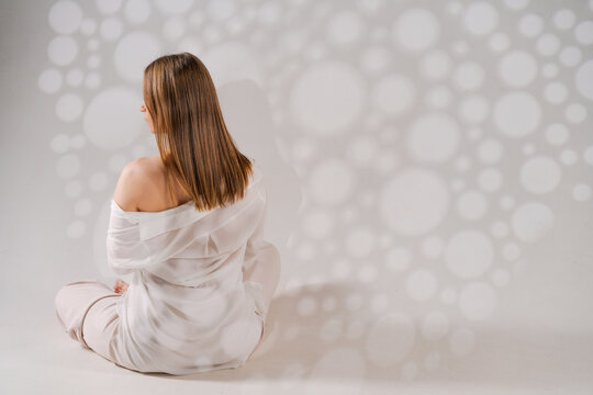 Depressed woman sits on floor on light background with soft, bright shadows, a sad Caucasian woman in light clothes turned her back in the image. Free from copy space.