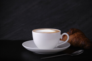 White cup with cappuccino on a dark background. Italian breakfast on a black background. Cappuccino with croissant on a black background.