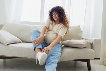Obraz premium Pregnant woman sitting on the couch at home and wearing comfortable shoes for pregnancy with a problem due to the stomach, headache, pregnancy and motherhood difficulties, severe fatigue