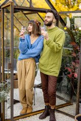 Young stylish couple talk and drink wine, spending leisure time together near glass house in...