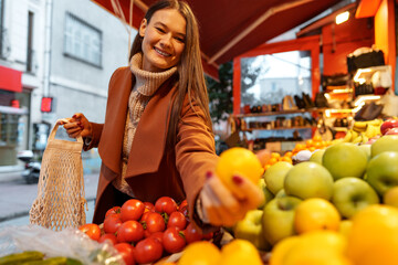 Good looking young woman standing in front of fruit shelves buying groceries in the street