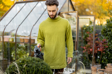Stylish guy takes care of plants spraying on on them in garden with glasshouse on background. Gardening hobby and topiary plants concept