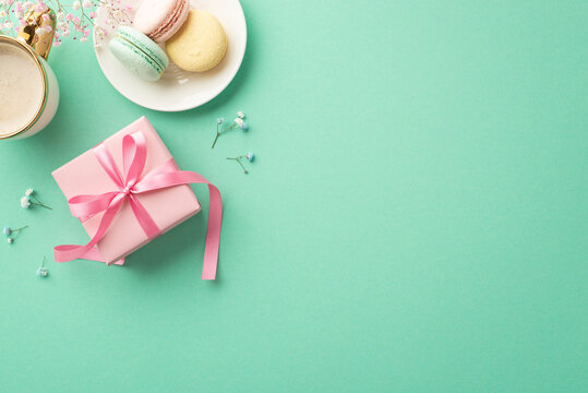 Spring presents concept. Top view photo of pink giftbox with ribbon bow plate with macarons cup of coffee and gypsophila flowers on isolated teal background with empty space