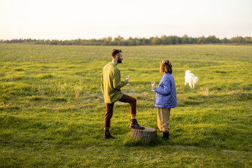 Young stylish couple hang out together, talking and drinking wine on green field during sunset. Spending autumn time outdoors