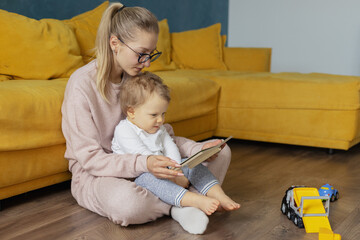 A young mother reads an interesting book to her baby at home on the floor