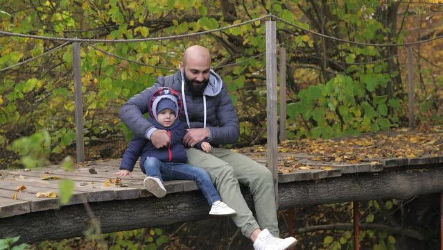 Bearded father and son are sitting on river bridge and looking at water flow. They talk, spend leisure time together. A man and 2 years old boy sit on a wooden bridge. Day vacation on river outdoors.