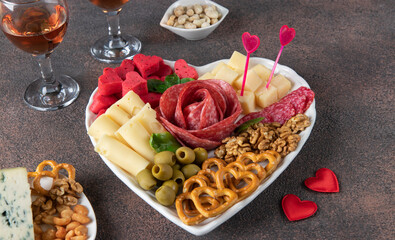 Plate shaped-heart for Valentines Day with varieties sausage, cheese, walnuts, olives and two glass rose wine on brown background