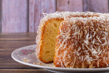 Coconut-flavored sweet cake topped with coconut flakes, with a cut slice showing the delicious dough on a decorated plate. Using wooden board and wooden background. Selective focus.