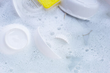 Dishes and bowls with yellow sponge in water and bubbles of dishwashing liquid