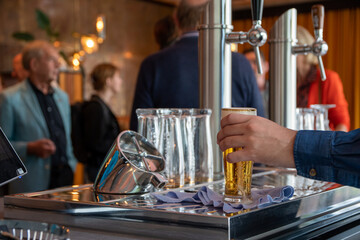 The hand of a caucasian bartender serving a glass of beer from behind his beer tap with people in the background