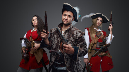 Shot of pirate man and two women with flintlock guns against grey background.