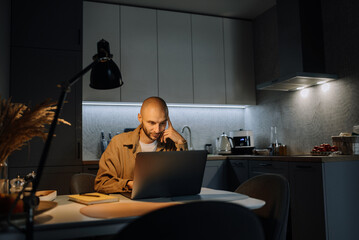 Stressed and worried man working at night on laptop computer at home in kitchen.