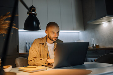A young man works on a laptop in a modern kitchen, checks e-mail in the evening, writes a message on a social network, a young man uses internet banking, searches for information